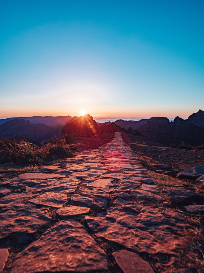 A long, empty stone pathway leading toward a powerful sunrise breaking behind majestic, sharp mountain peaks, symbolizing self-mastery and internal focus.