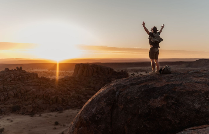 A solitary person standing triumphantly on a rocky cliff silhouette facing a massive golden sunrise over a vast desert landscape.