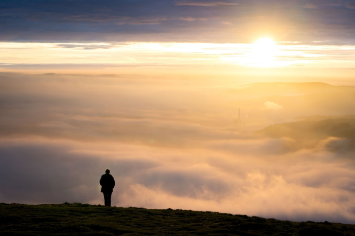 A lone man standing on a rocky mountain summit, looking out over a thick sea of clouds illuminated by warm golden sunrise light.
